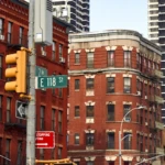 Street corner with red brick buildings, traffic lights, and various signs including "Wrong Way" and "E 118 St." Urban setting with a clear sky.
